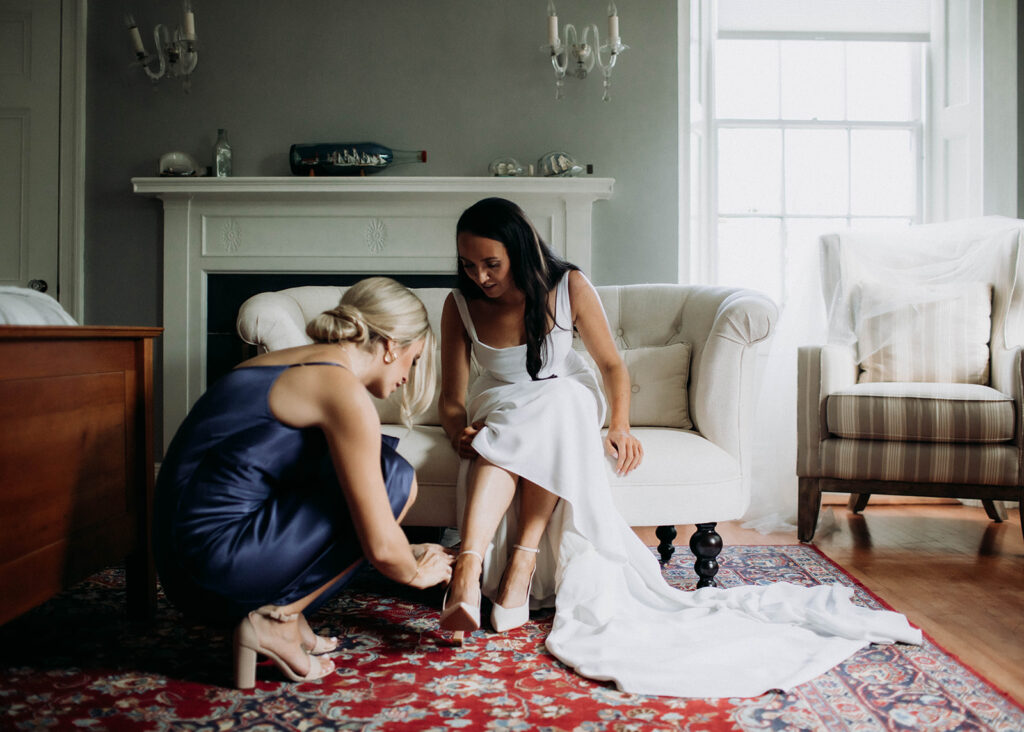 A women in a wedding dress is sitting a couch while her friend crouches on the floor helping her put her shoes on. This is a photo from a romantic coastal wedding in St. Andrews, New Brunswick photographed by Shannon-May