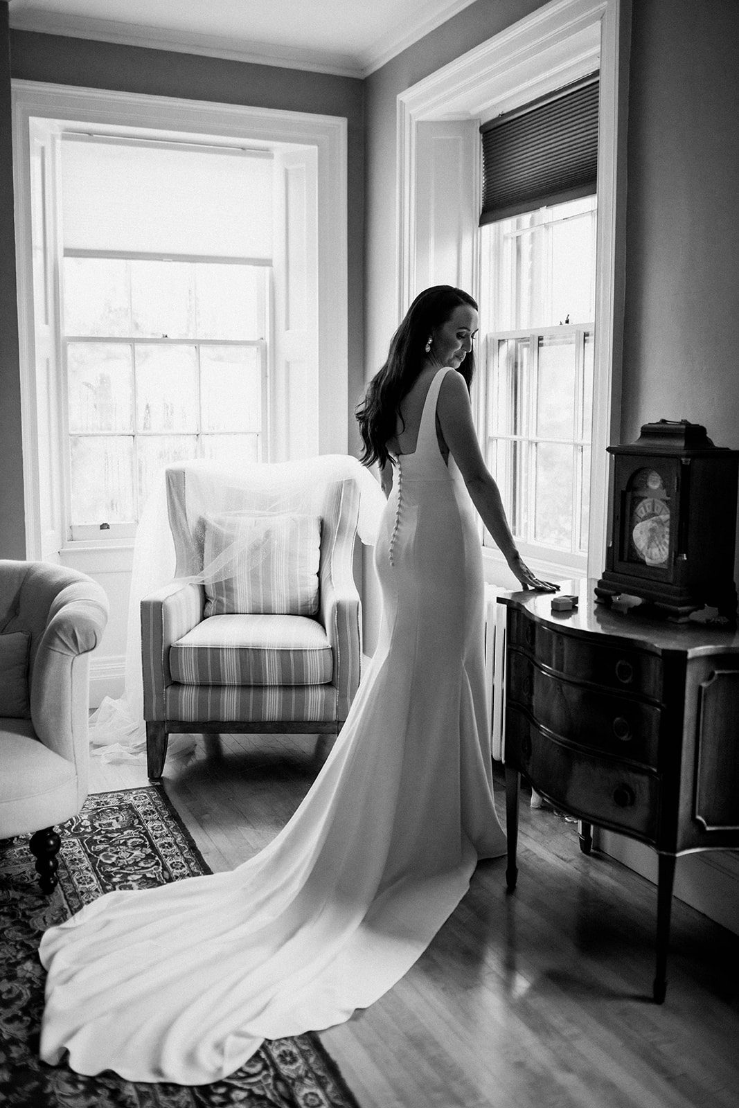 A women in a wedding dress stands towards a window. Her right hand is on a table and she is looking down at her right hand. Her wedding dress is long and spread out behind her. Her dress is laid across the back of a chair in the background. This is a photo from a romantic coastal wedding in St. Andrews, New Brunswick photographed by Shannon-May