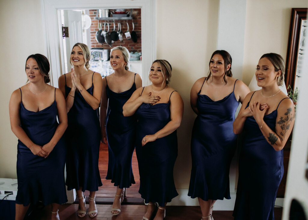 A group of 6 bridesmaids in blue midi dresses stand looking up with smiles and tears in their eyes. They are looking at their friend (not pictured). This is a photo from a romantic coastal wedding in St. Andrews, New Brunswick photographed by Shannon-May.