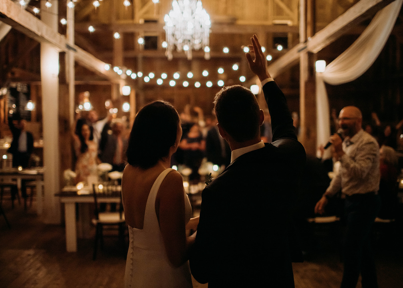 a newly married couple stand holding hands with their backs to the camera looking towards their wedding guests and the man's arm is raised and waving.