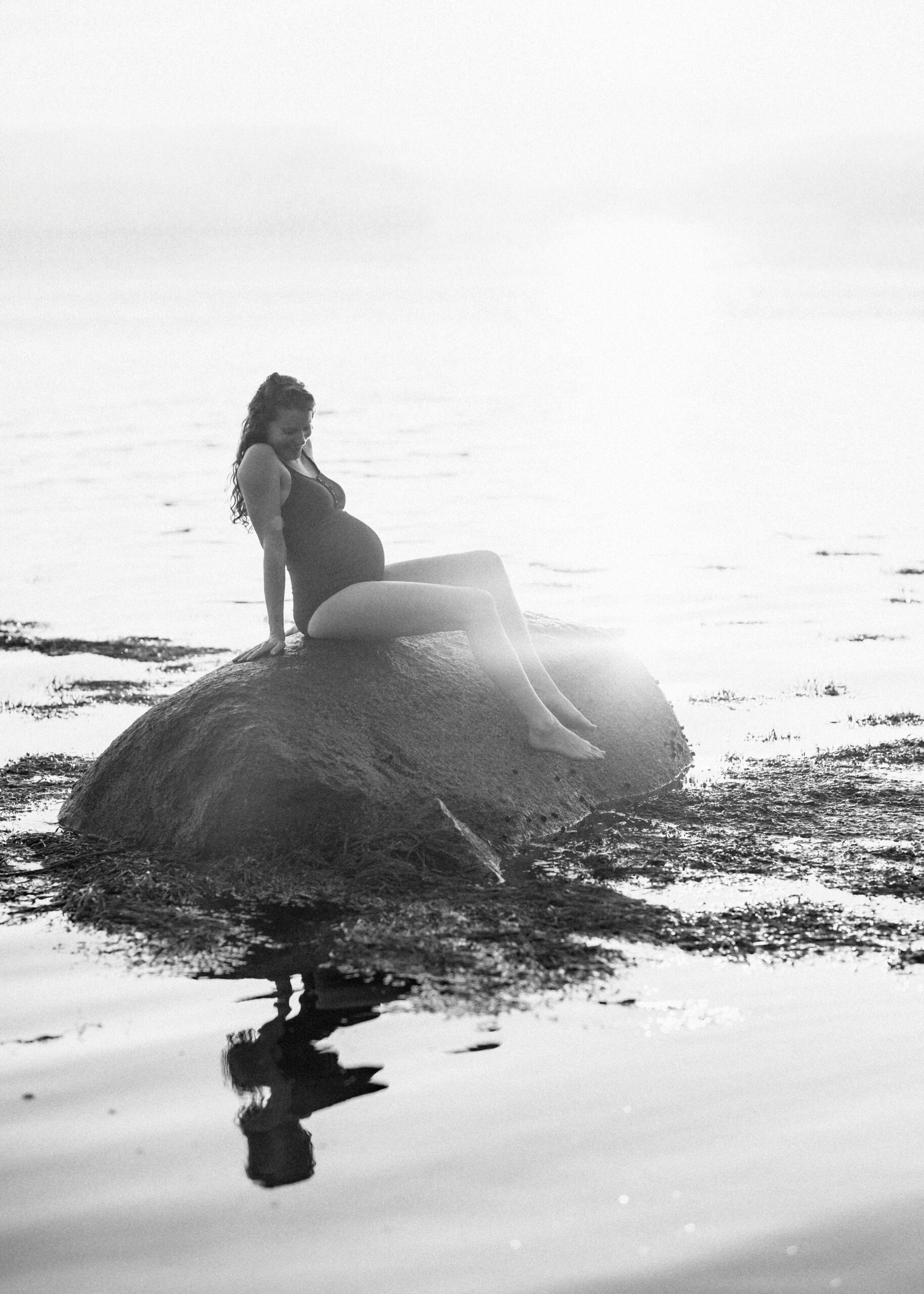 A black and white image of a pregnant woman sitting on a large rock in the ocean. She leans back showing off her pregnant belly. 