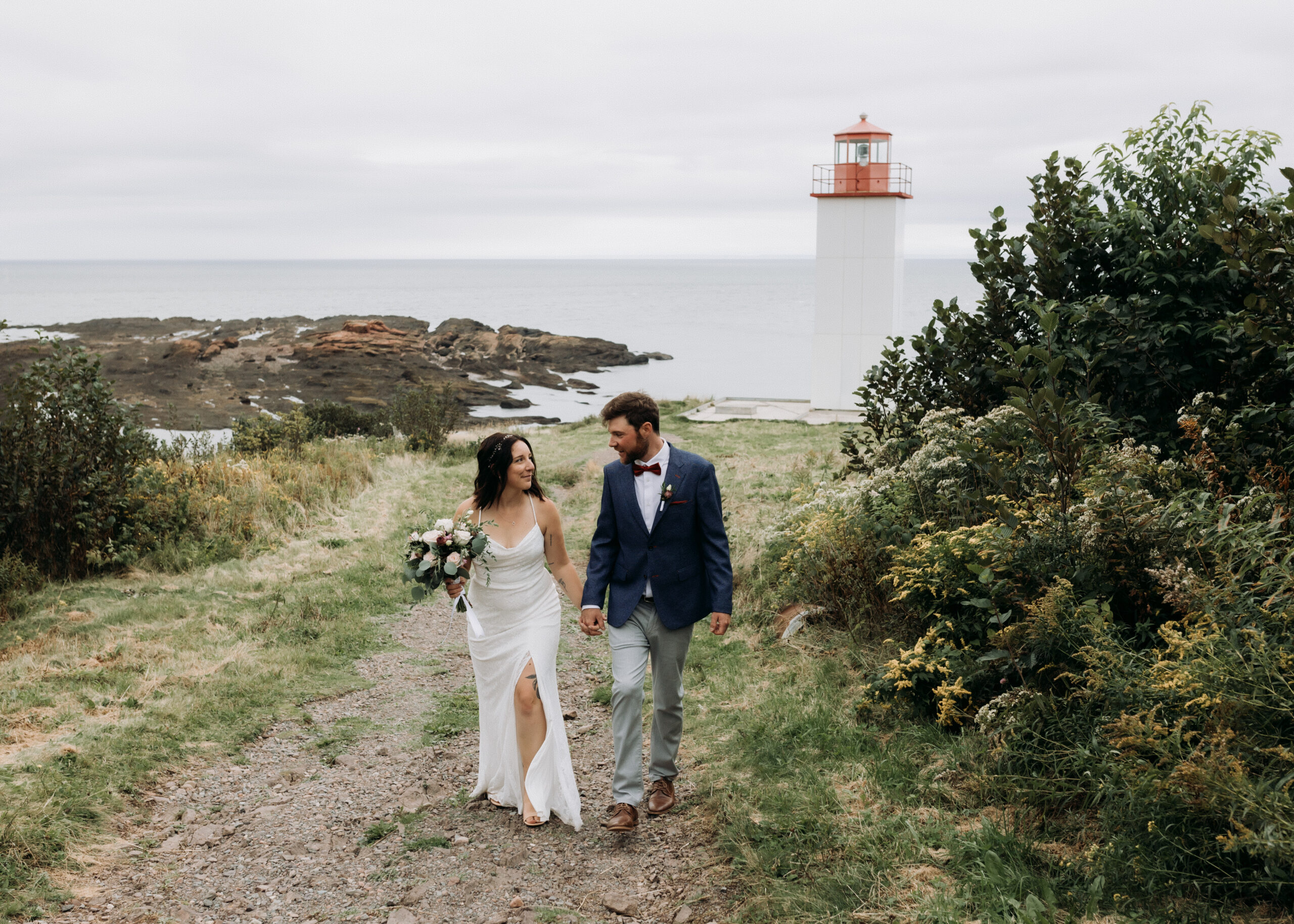 elope in new brunswick. a newly married couple walks hand in hand towards the camera. the ocean and a lighthouse are behind them in the background.