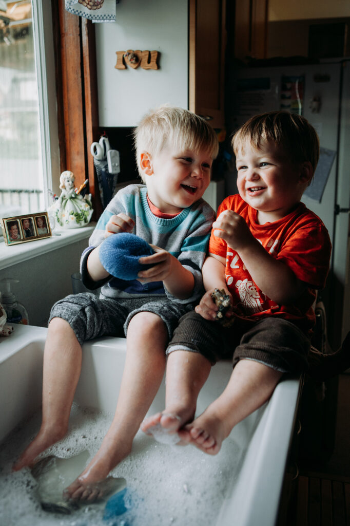 two little boys sit in their grandmother's kitchen sink having their little feet washed. this is the perfect example of why photographing families in their home is so beautiful.