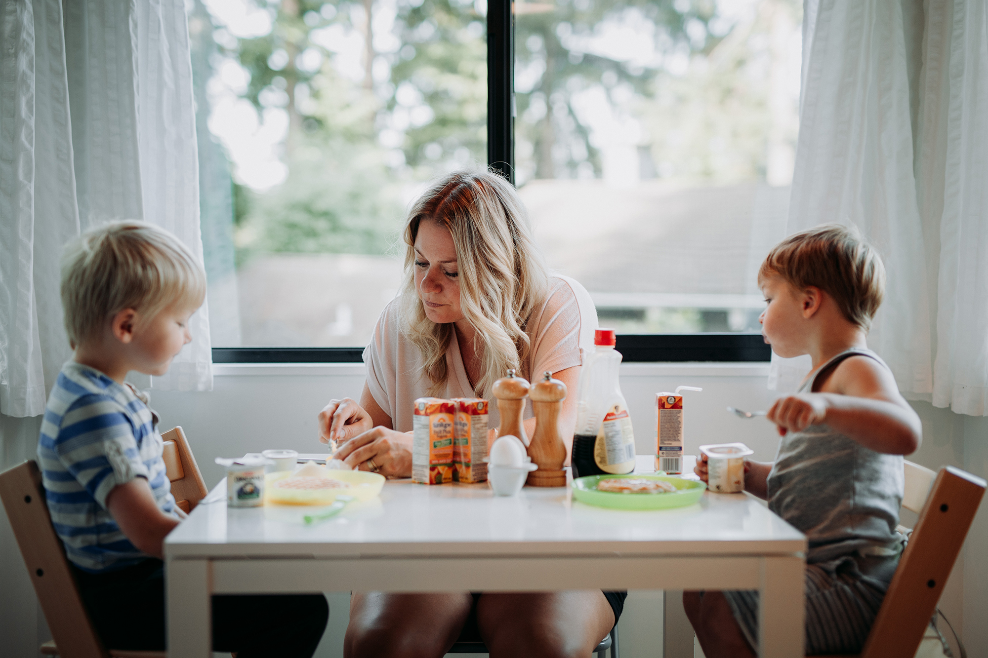 a mother sits a small kitchen table with her two young sons. she is cutting up a piece of fruit while her little boys look on with wonder and curiousity.